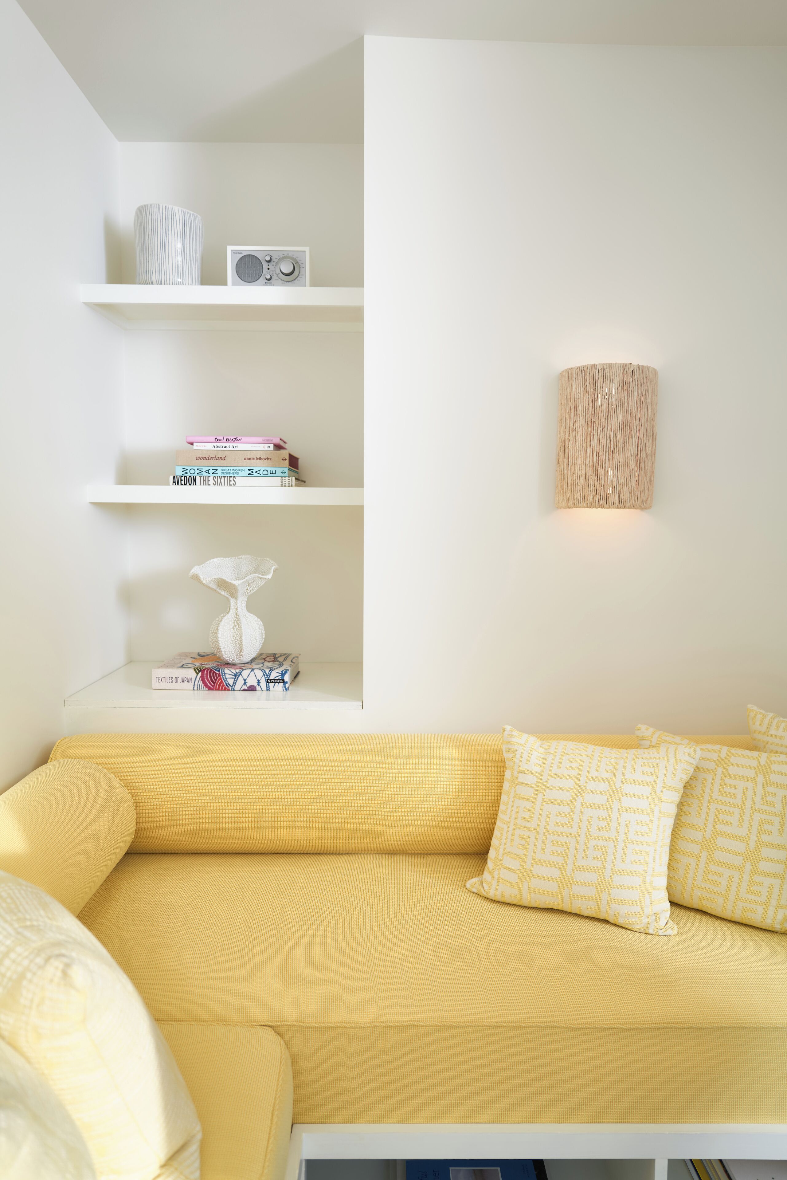 Una sala de estar moderna con un sofá amarillo, estanterías blancas con libros y objetos decorativos, y una lámpara de pared con textura.