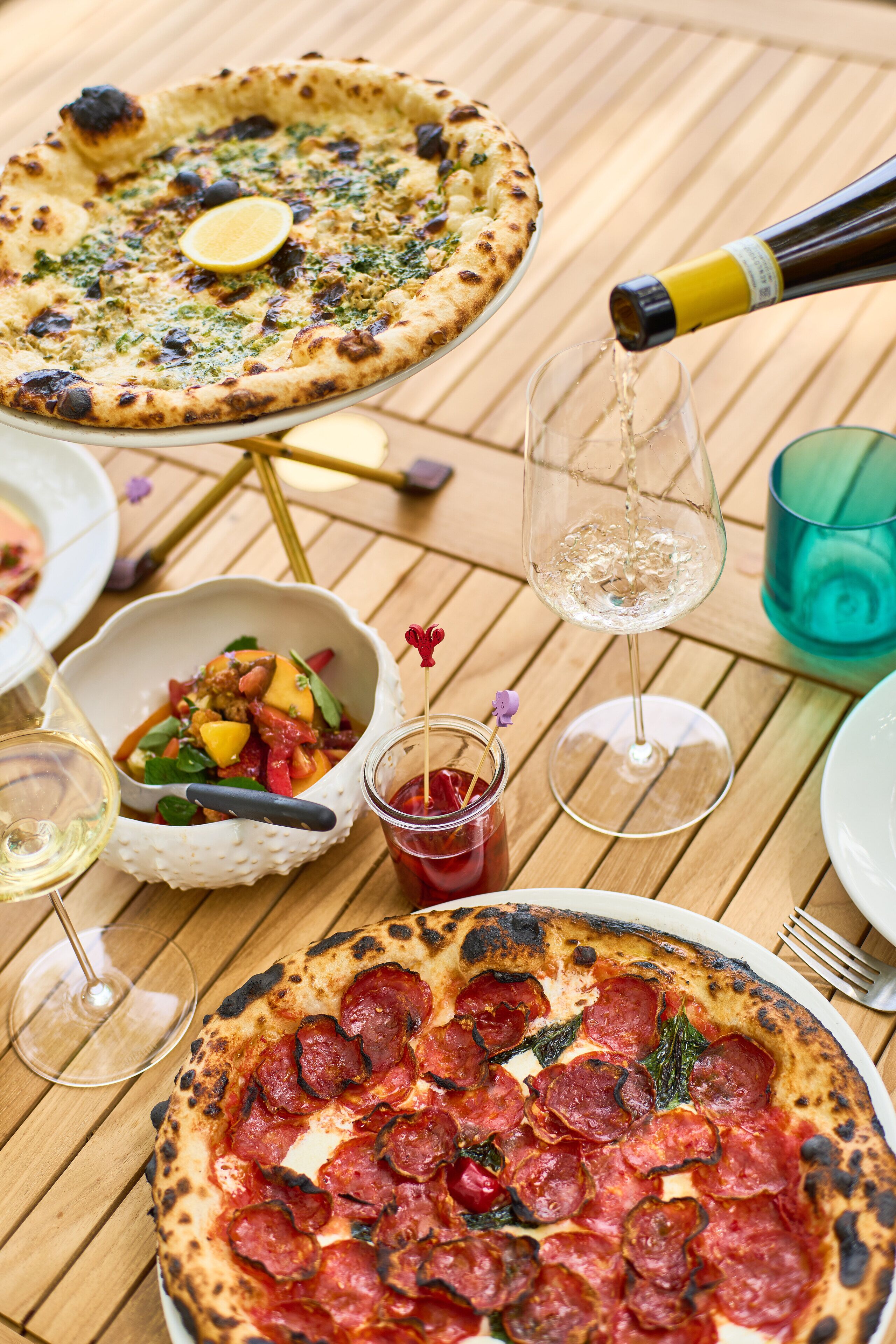 Zwei Pizzen auf einem Holztisch, während Wein in ein Glas gegossen wird.