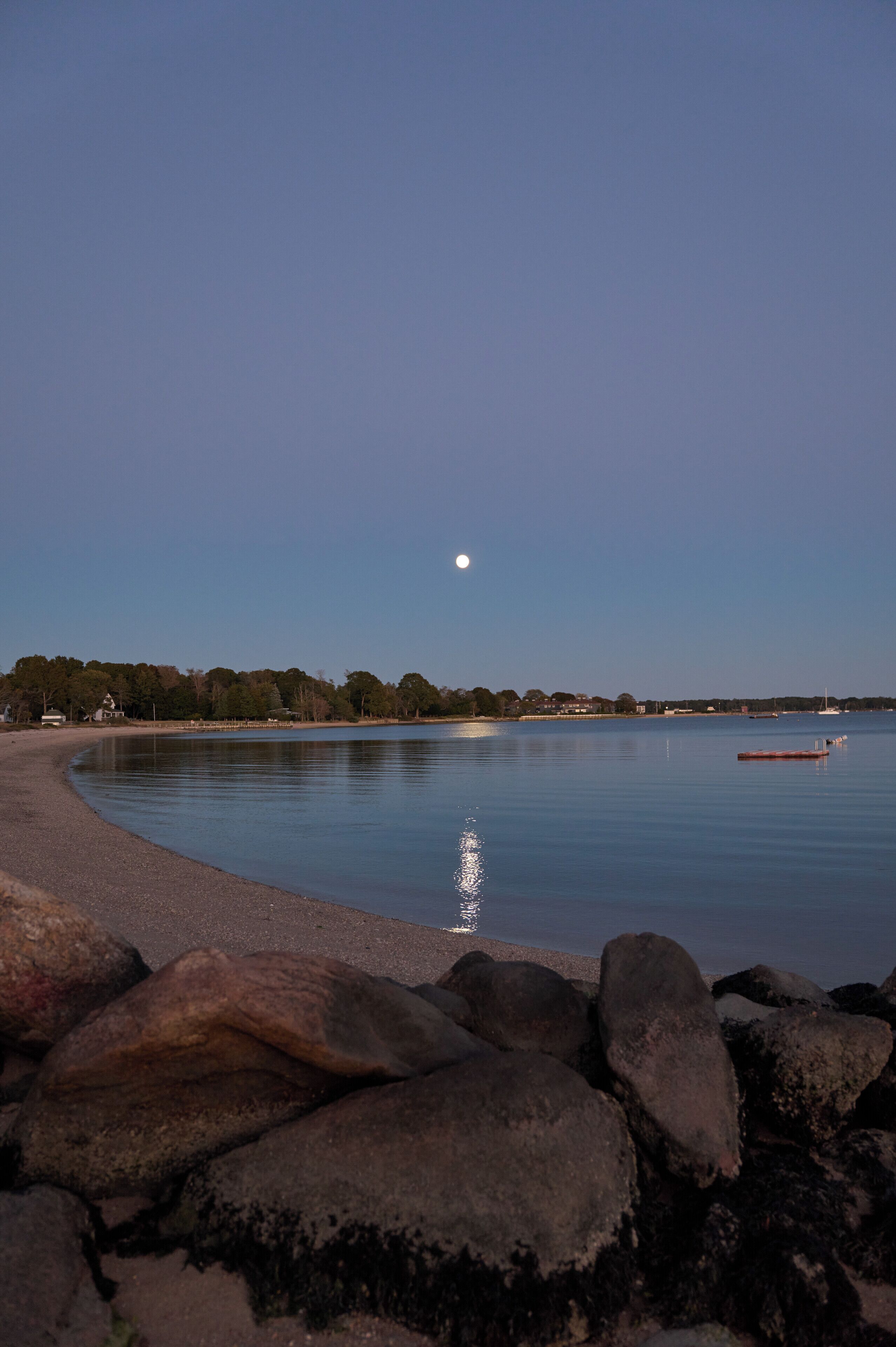Um lago tranquilo ao entardecer com uma lua visível, cercado por pedras e árvores.