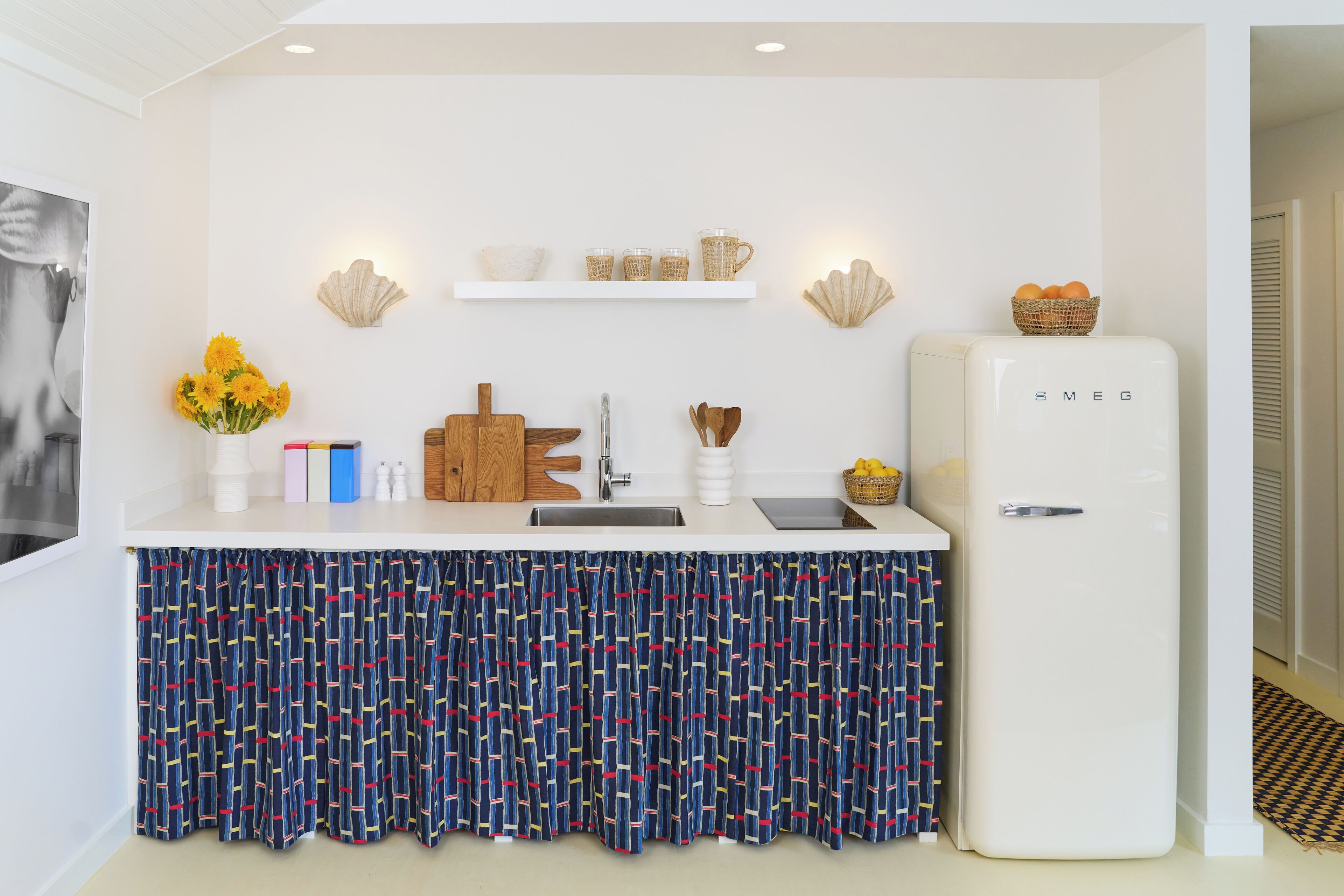 Une cuisine de taille réduite avec un réfrigérateur blanc, un évier et un plan de travail. On peut y voir des coquillages décoratifs et un panier accrochés au mur, ainsi qu’un vase rempli de fleurs posé sur le plan de travail.