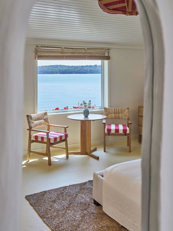 Una habitación con una cama, dos sillones de mimbre con cojines de cuadros, una pequeña mesa redonda con un florero y vistas a un lago a través de una ventana con persiana de bambú.