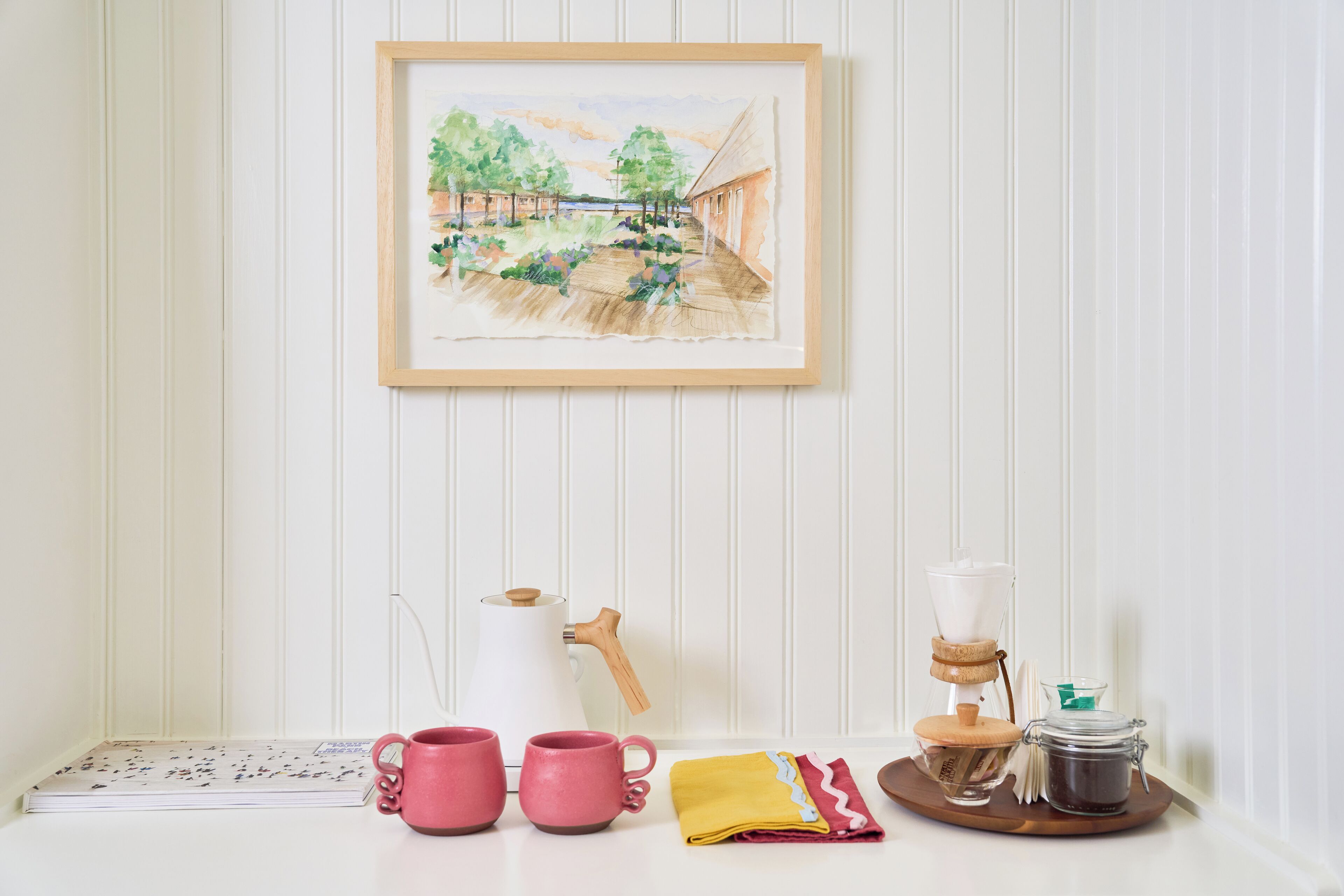 Un tavolo ordinato con un quadro acquerellato incorniciato, una macchina per il caffè, tazze rosa, un tovagliolo giallo e un barattolo di chicchi di caffè.