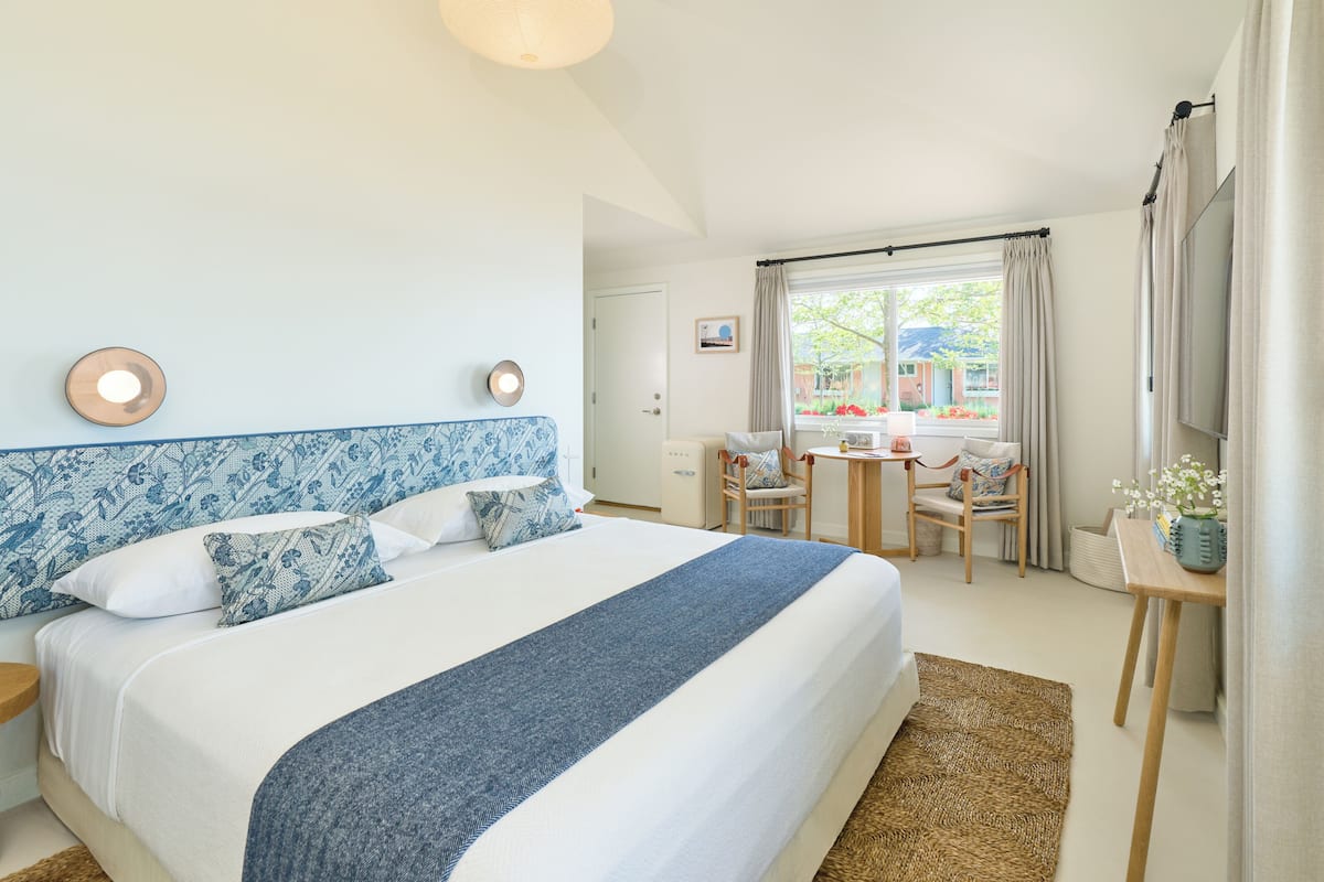 A bedroom with a large bed, a desk, and a chair. There is a window with curtains and a view of the outside.