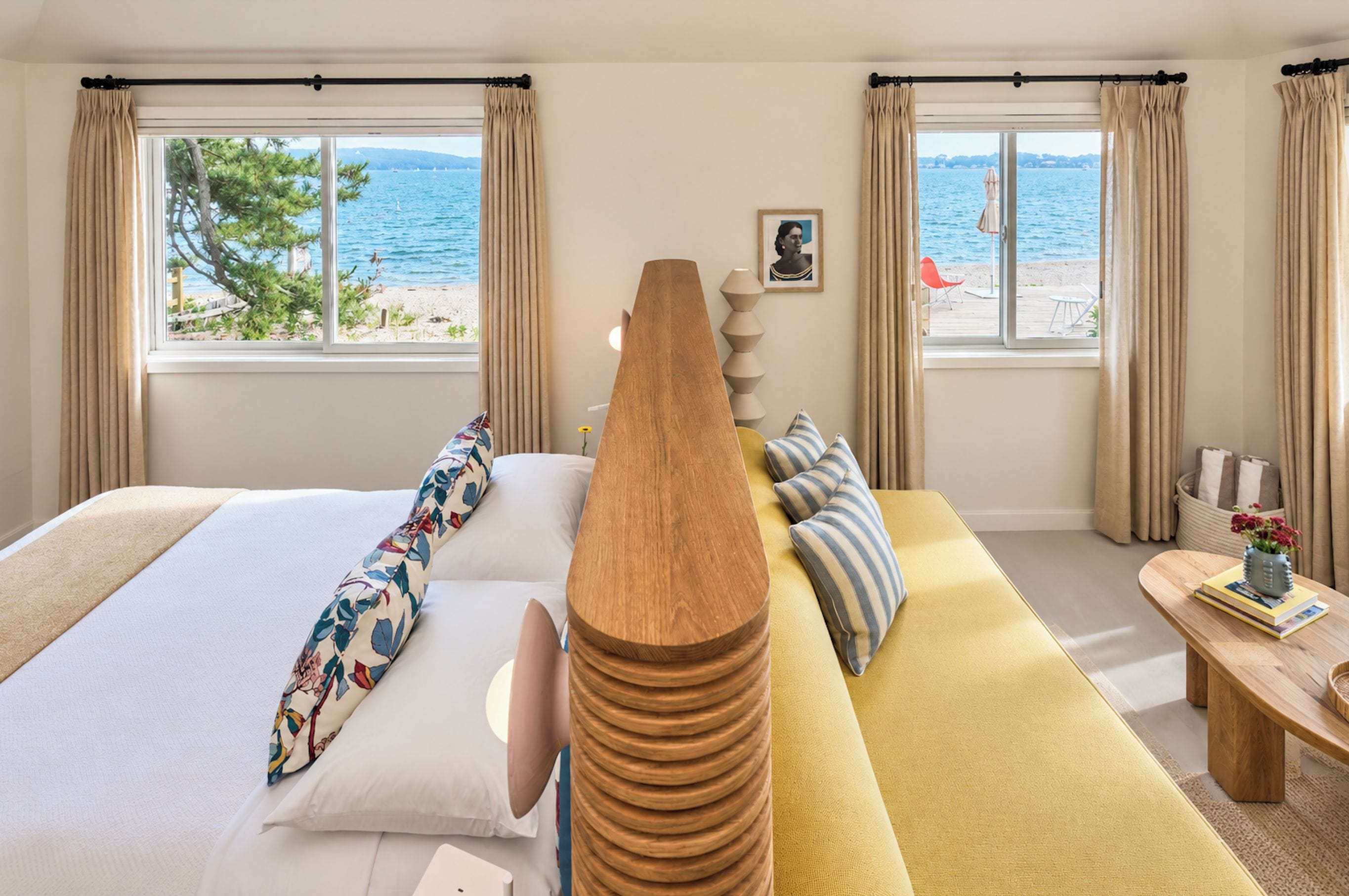 A hotel room with a bed, a wooden headboard, a sofa, a small table with a vase, and a view of the beach and sea through the windows.