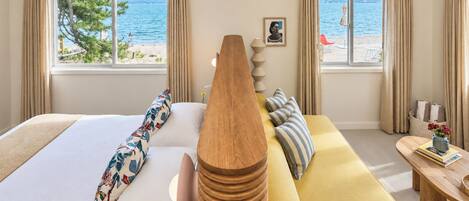 A hotel room with a bed, a wooden headboard, a sofa, a small table with a vase, and a view of the beach and sea through the windows.
