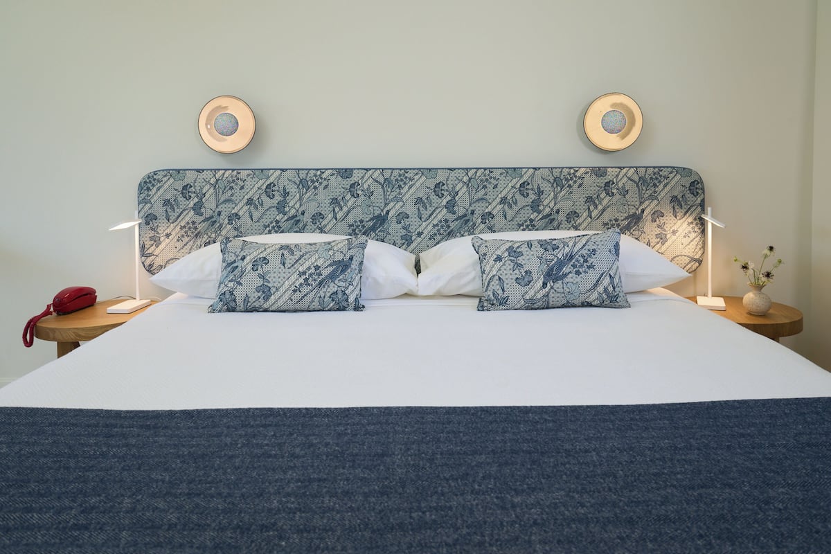 A neatly made bed with patterned pillows and a blue headboard, two bedside lamps, a red telephone, and a small vase with flowers on a nightstand.