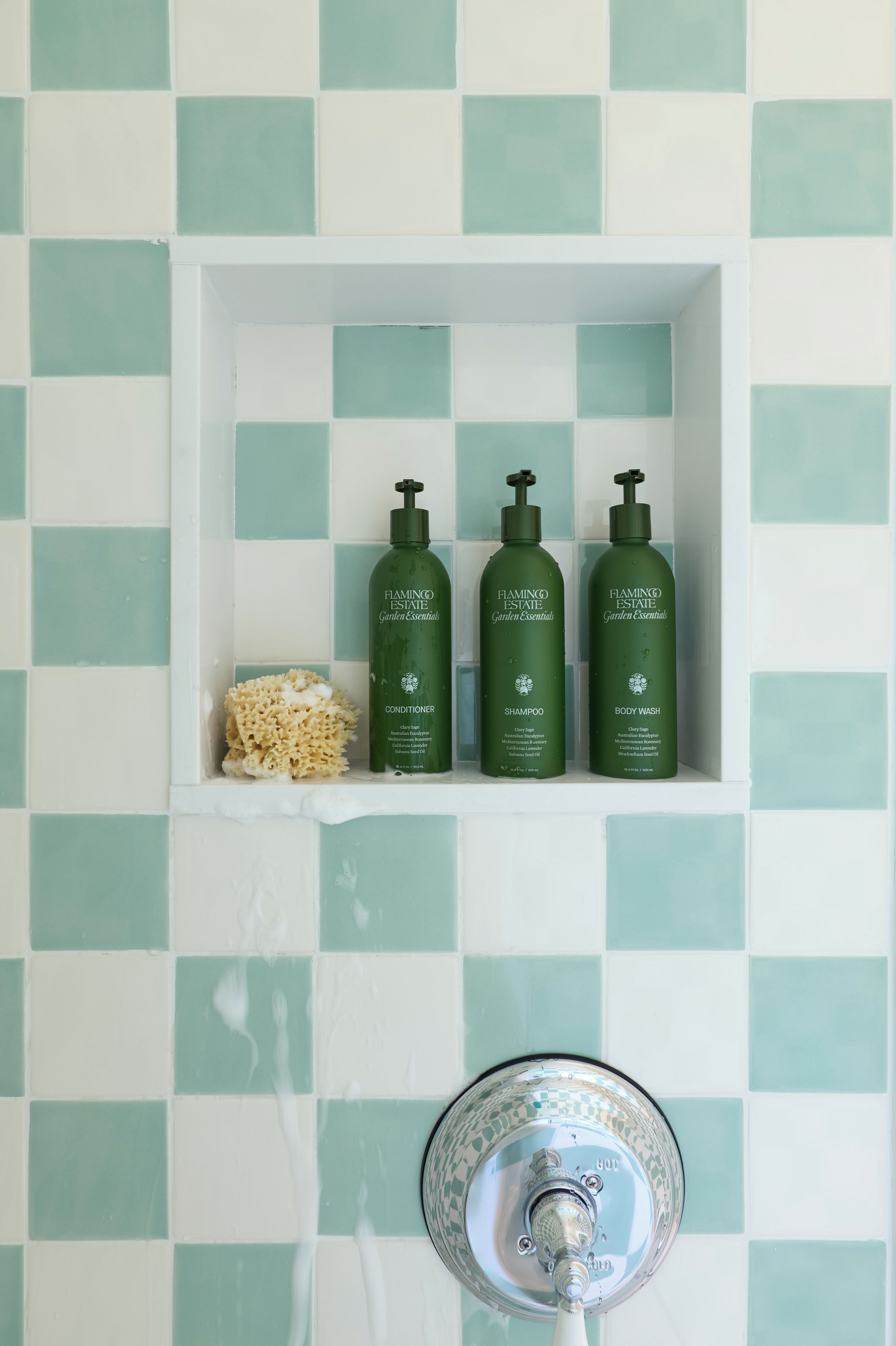 Une salle de bain avec un mur recouvert de carreaux à motifs, une étagère sur laquelle sont posées trois bouteilles vertes étiquetées « Indispensables du jardin de Flamingo Estate », et une pomme de douche.