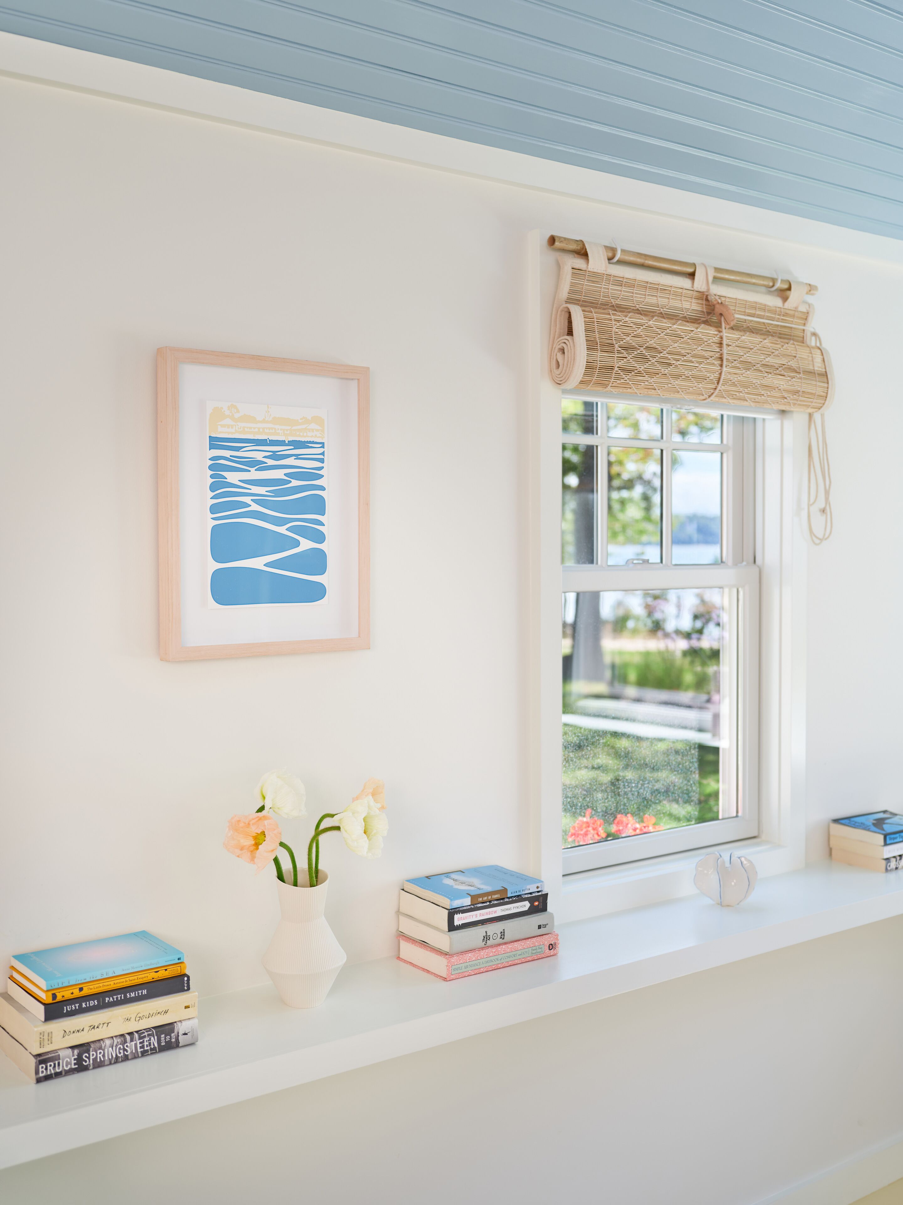 Ein Zimmer mit weißer Wand, einem gerahmten Kunstwerk, einem Fenster mit Bambusrollvorhang, einer Vase mit Blumen und einem Bücherstapel auf einem Regal.