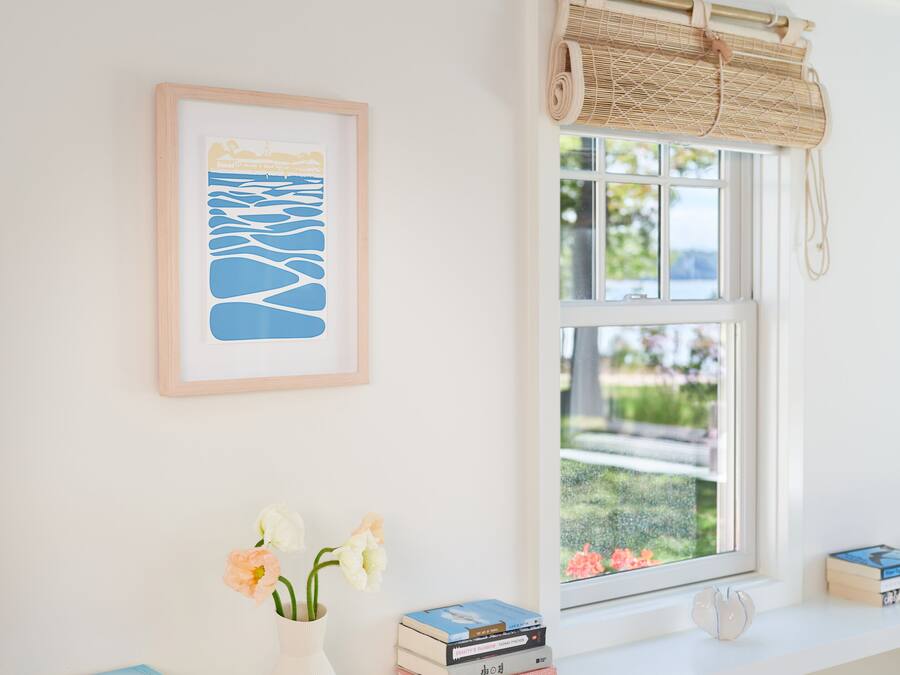Ein Zimmer mit weißer Wand, einem gerahmten Kunstwerk, einem Fenster mit Bambusrollvorhang, einer Vase mit Blumen und einem Bücherstapel auf einem Regal.