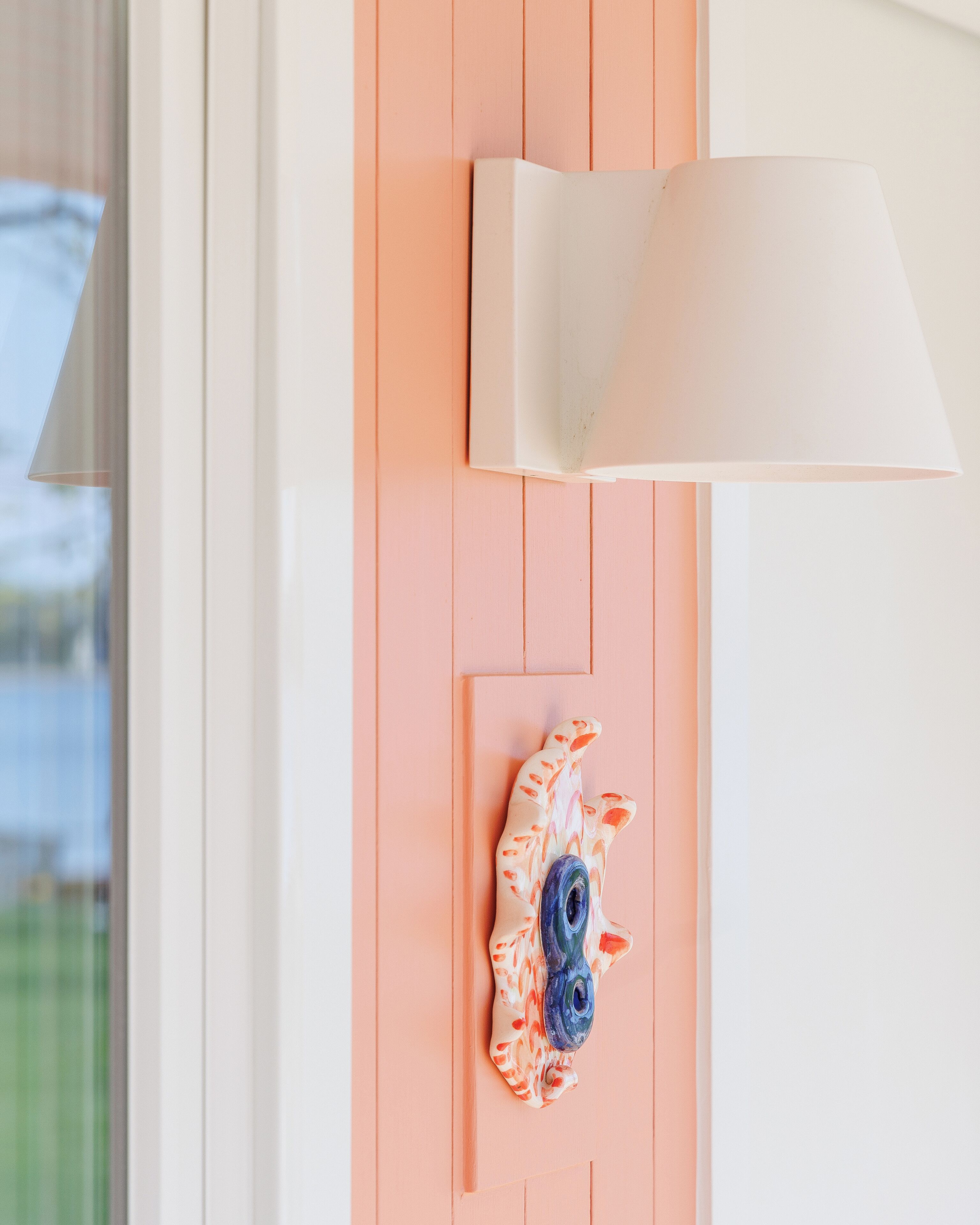A wall-mounted lamp with a white shade, a decorative ceramic piece, and a door with a peach-colored panel.