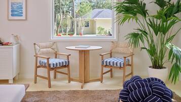 A room with two chairs, a small table, a large potted plant, and a window with a view of trees and a house.