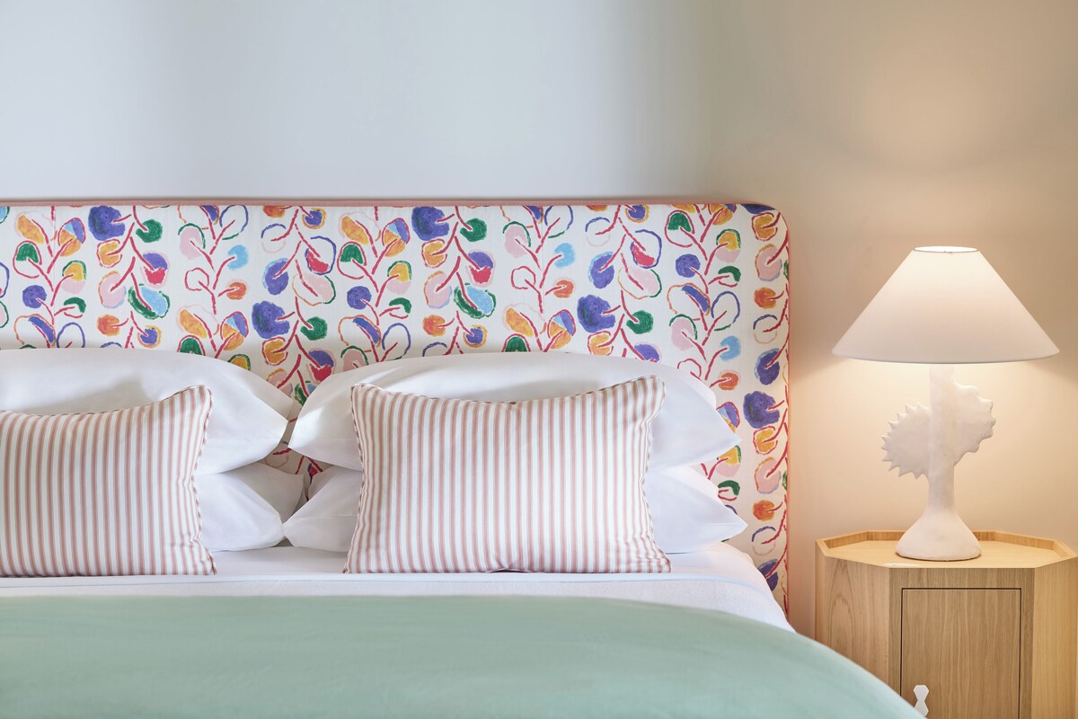 Una cama con almohadas a rayas y un cabecero colorido, una lámpara de noche y una mesita de madera.