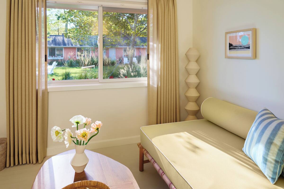 A room with a bed, a window with curtains, a vase with flowers, and a framed picture on the wall.
