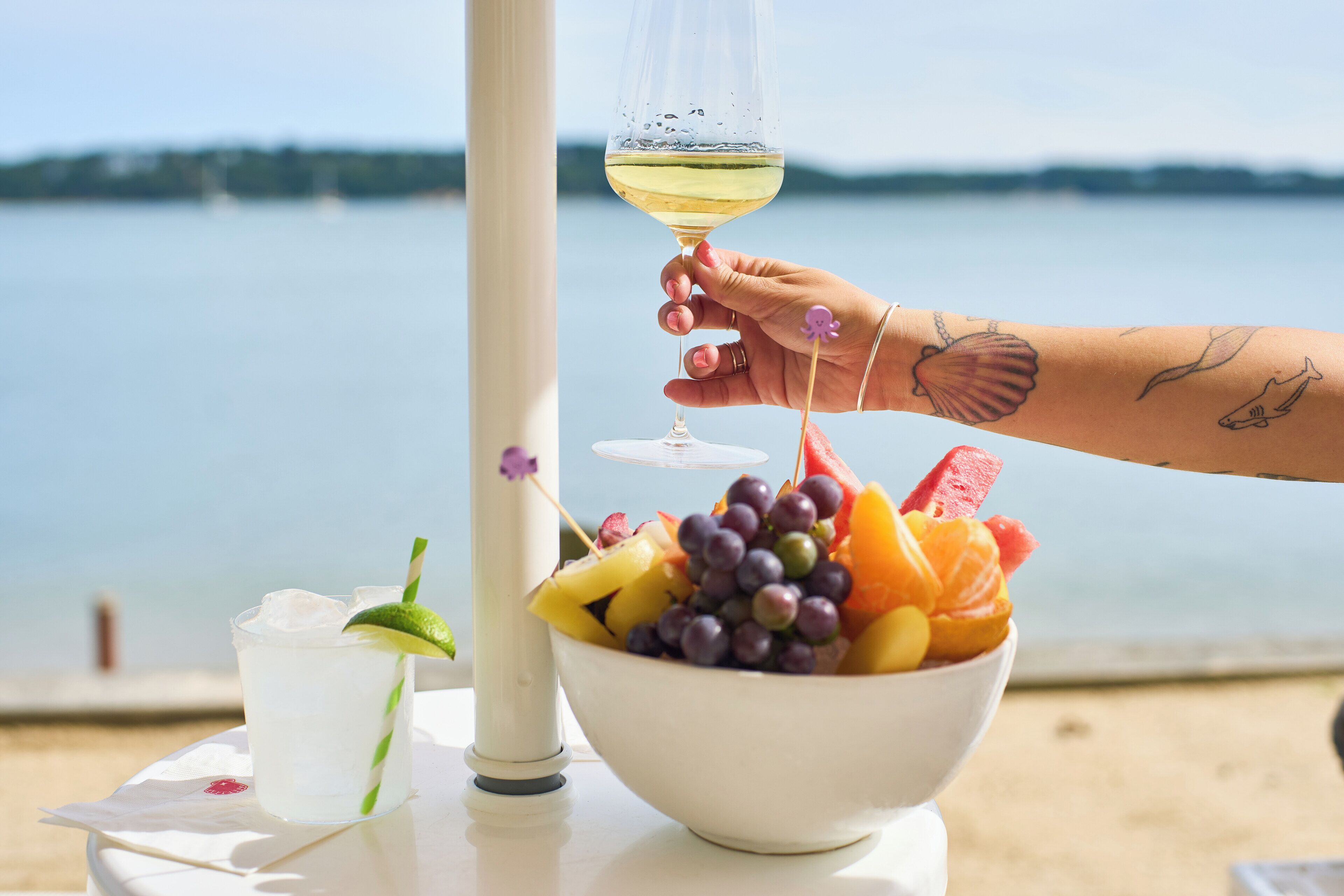 A table with a glass of white wine, a bowl of fruit, and a drink with a lime wedge.