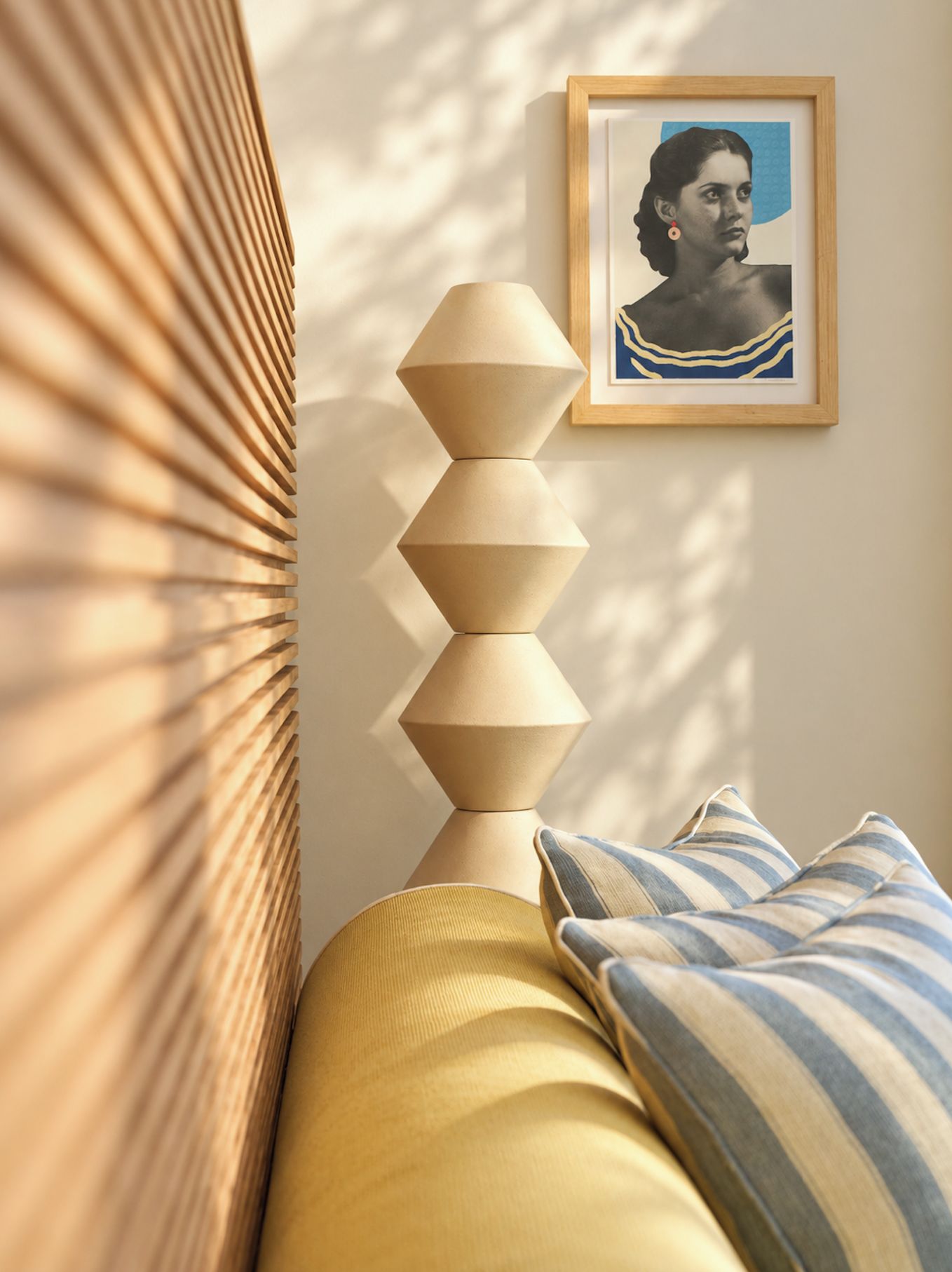 Una cama con almohadas dispuestas geométricamente, una obra de arte enmarcada en la pared y una ventana con persianas proyectando sombras.