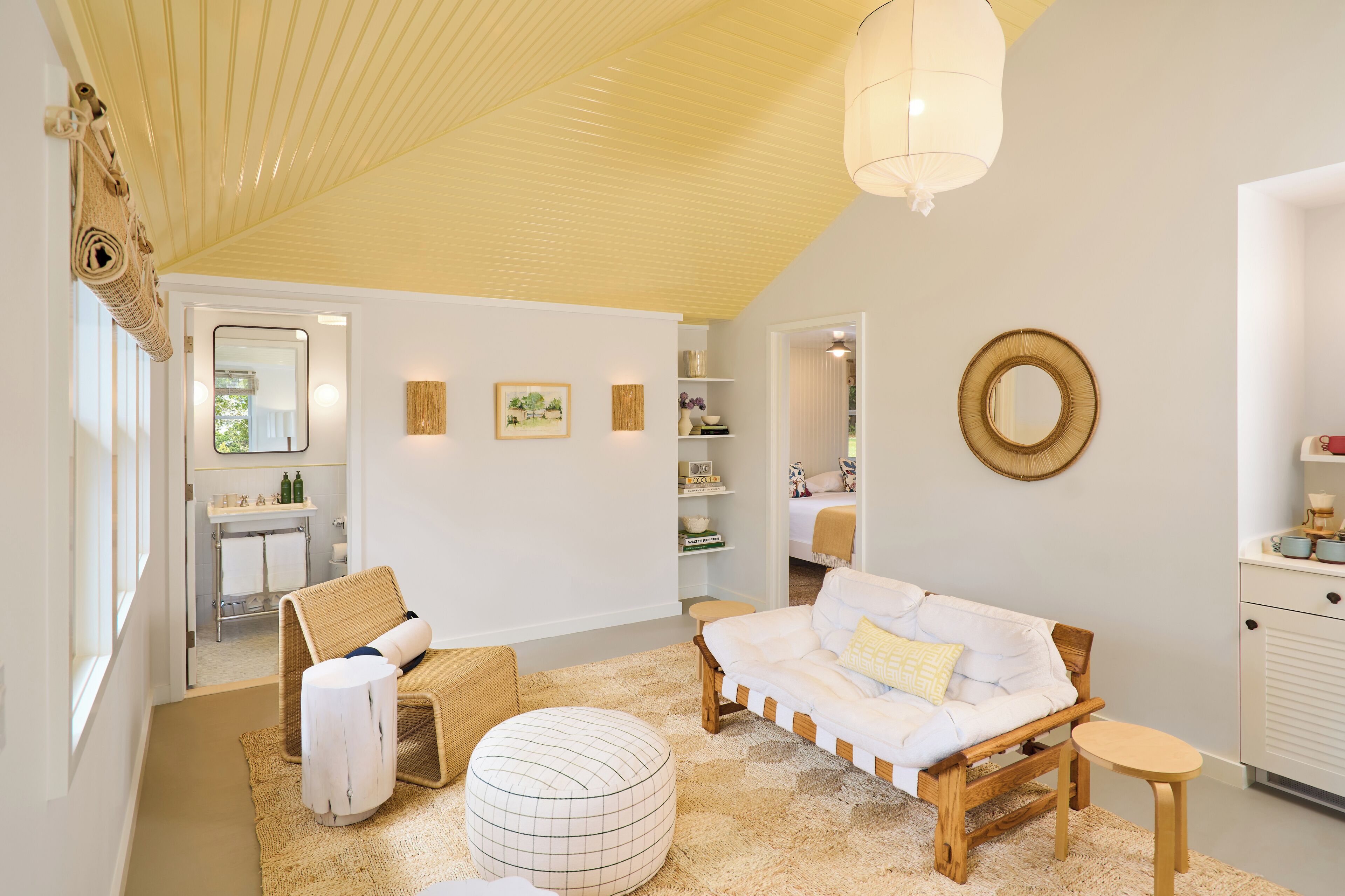 Quarto aconchegante com um banco de madeira, um sofá branco e uma mesa pequena. Há um espelho na parede e uma prateleira com objetos decorativos.