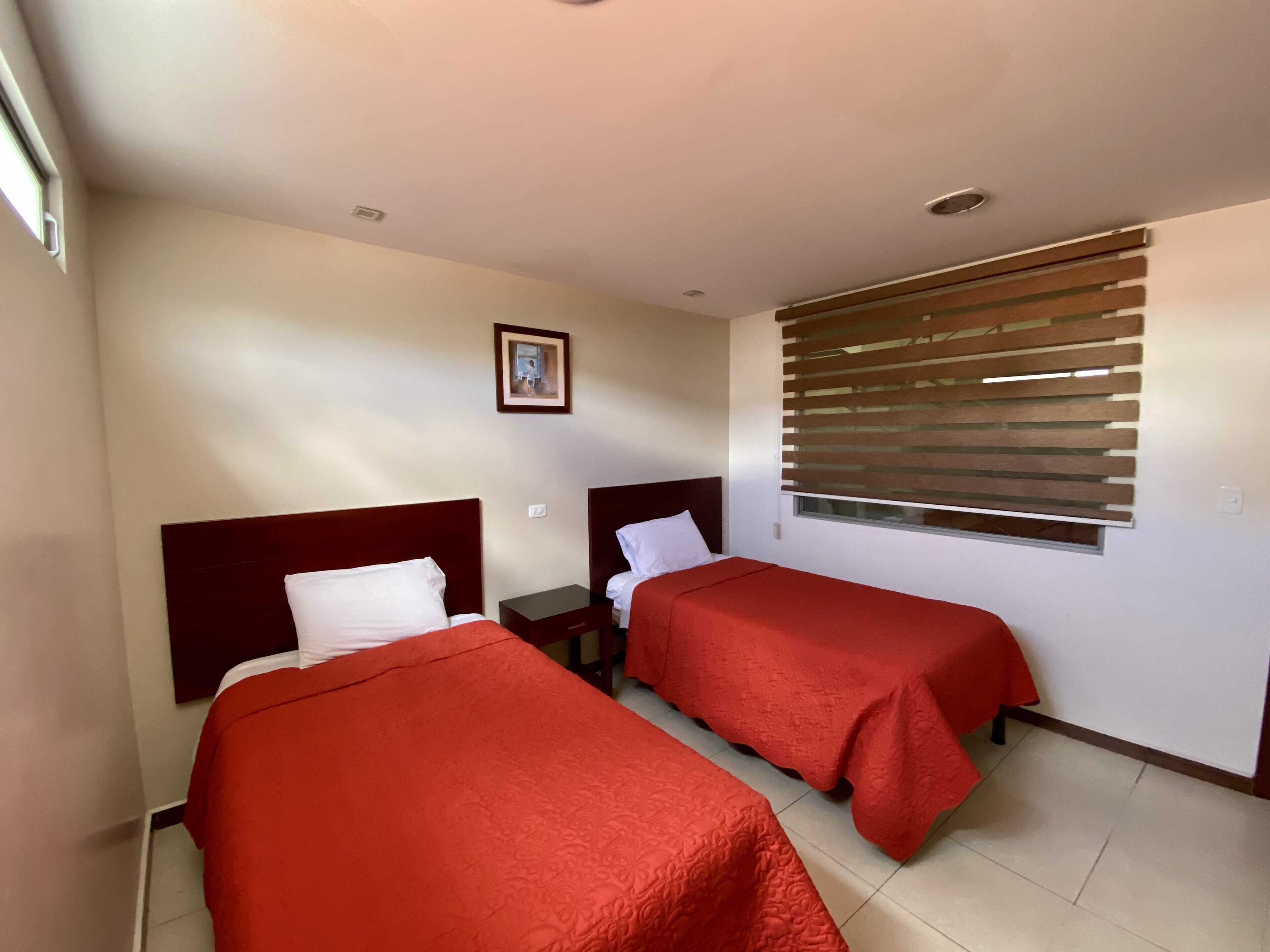 Quarto de hotel com duas camas, janela com persianas, quadro na parede e uma pequena mesa de cabeceira.