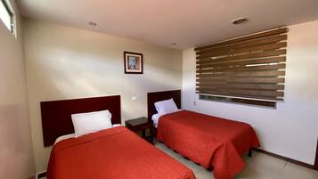 Quarto de hotel com duas camas, janela com persianas, quadro na parede e uma pequena mesa de cabeceira.