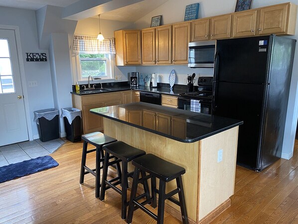 A modern kitchen with wooden cabinets, a black refrigerator, a microwave, and a large island with bar stools. - Enjoy the Best of Both Worlds on Narrow River and Narragansett Beach (South Kingstown)