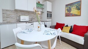 A modern kitchen with white cabinets, a built-in oven, and a microwave. There is a dining table with chairs and a vase with flowers. A gray sofa with red and yellow cushions is in the living area.