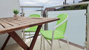 A wooden table with a potted plant, two green chairs, and a white chair on a balcony with a view of a hill and buildings.
