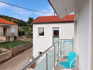 Un balcon avec une chaise bleue, une petite fenêtre et une vue sur une route et des bâtiments en arrière-plan.