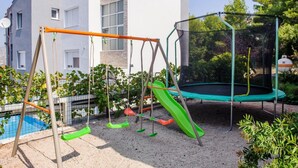 Un’area giochi con altalene, scivolo e trampolino, immersa nel verde e adiacente a una piscina.