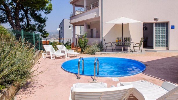 Area piscina con sedie a sdraio e un tavolo, circondata da un edificio con balconi e vista sul paesaggio.