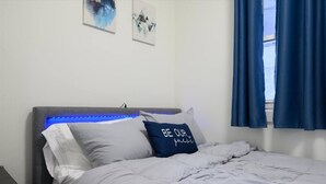 Una cama bien hecha con una almohada que dice 'BE OUR guest', una tira de luz LED azul debajo del cabecero y dos obras de arte enmarcadas en la pared.