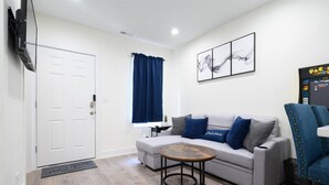 Una sala de estar con un sofá, una mesa de centro, una silla, un televisor, una puerta, una ventana con cortinas y obras de arte en la pared.