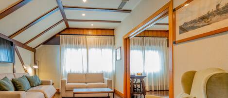 A cozy living room with a sofa, a coffee table, and a chair. There is a wooden ceiling with exposed beams, and a painting on the wall.