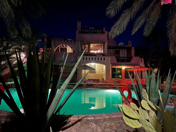 A nighttime view of a hotel with a swimming pool, palm trees, and a lit-up building.