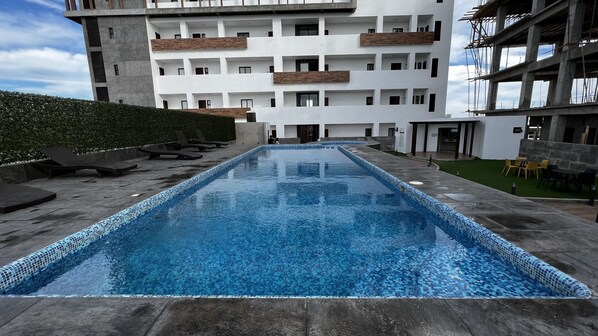 Una piscina amb vora de rajola, envoltada per un passeig pavimentat, i un edifici amb diversos balcons al fons.
