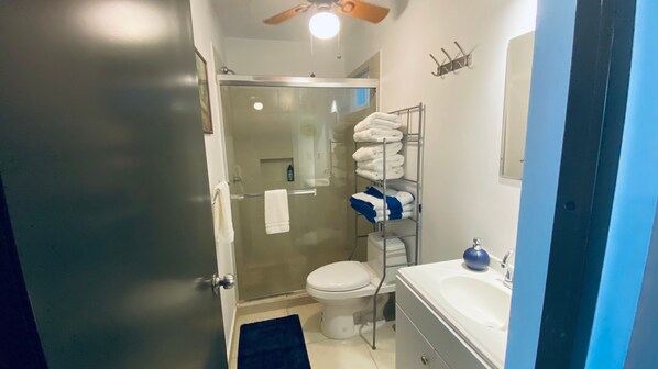 Un baño con cabina de ducha de vidrio, un inodoro blanco, un lavamanos con dispensador de jabón azul y un perchero con toallas dobladas con cuidado.