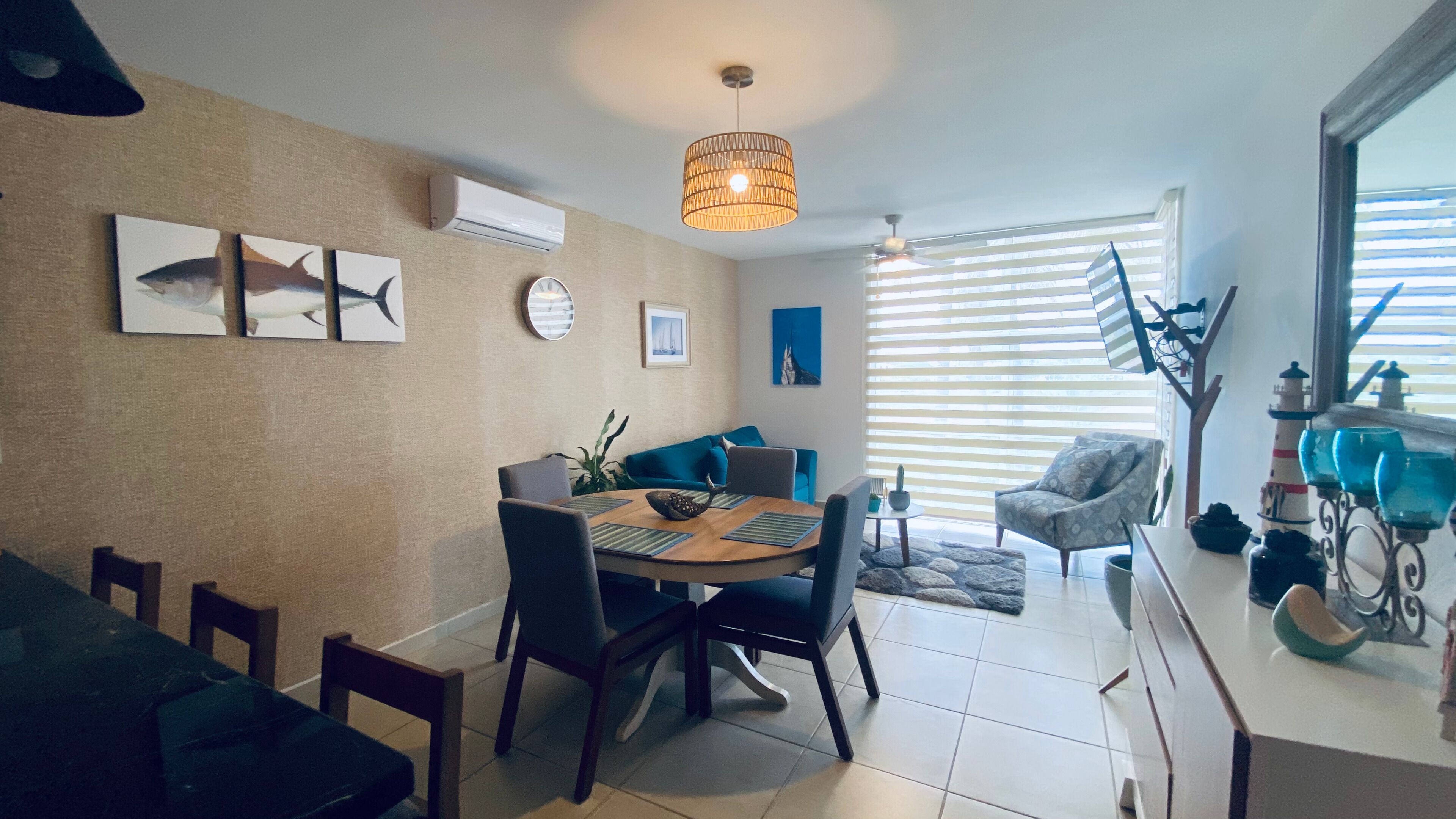 Een eetruimte met een ronde tafel, stoelen en een plafondventilator. Er hangen ingelijste foto’s aan de muur en er is een raam met zonwering.