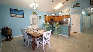 A dining area with a wooden table and white chairs, a kitchen with wooden cabinets and stainless steel appliances, and a blue wall with decorative items. - Welcome to Casa del Mar! Less than a mile away from beach! (Port Aransas)