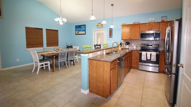 A kitchen with wooden cabinets, a stainless steel refrigerator, and a dining area with a table and chairs. - Welcome to Casa del Mar! Less than a mile away from beach! (Port Aransas)