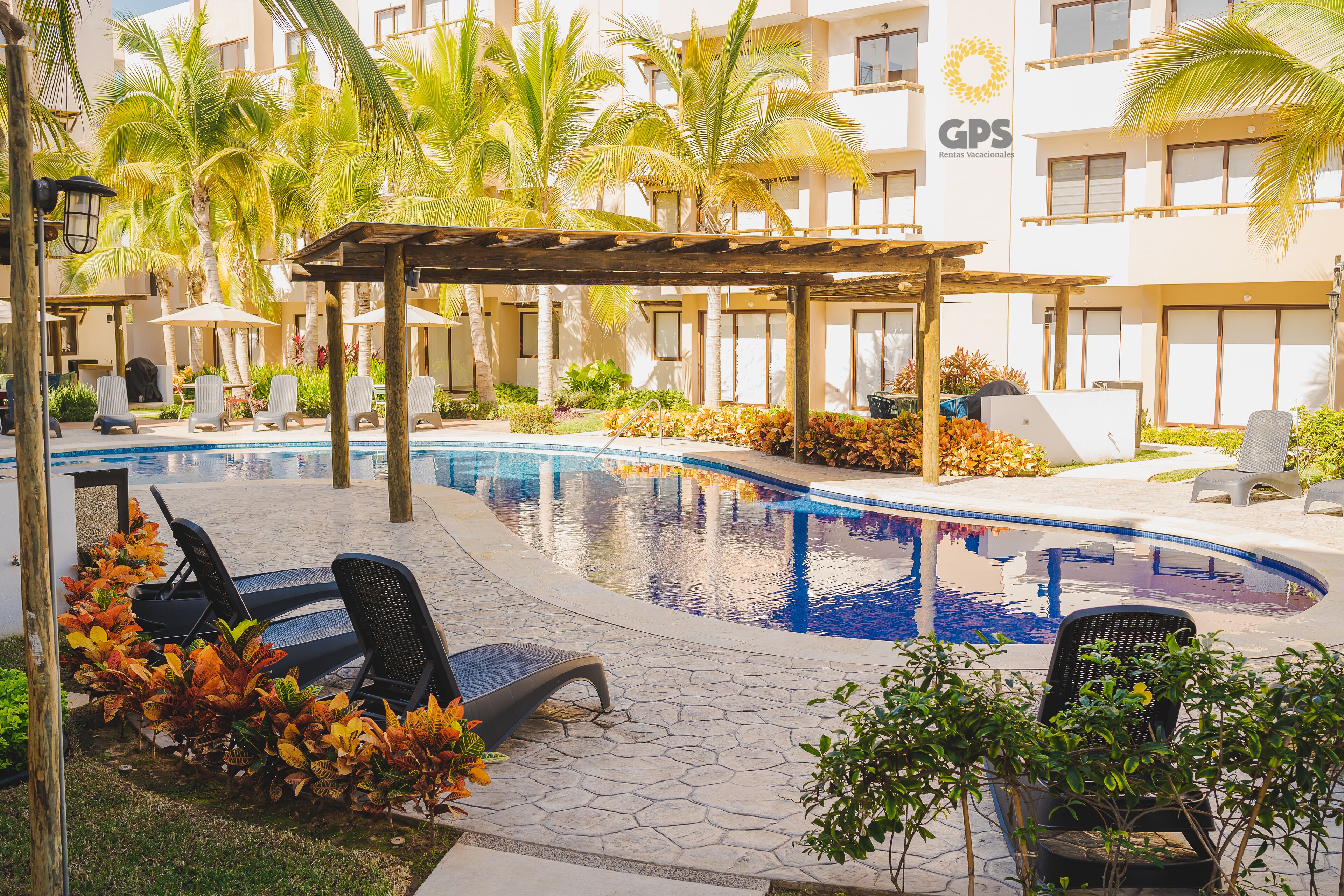 Zona de piscina con sillones de descanso, palmeras y un edificio con un cartel que dice 'GPS Rentas Vacacionales'.