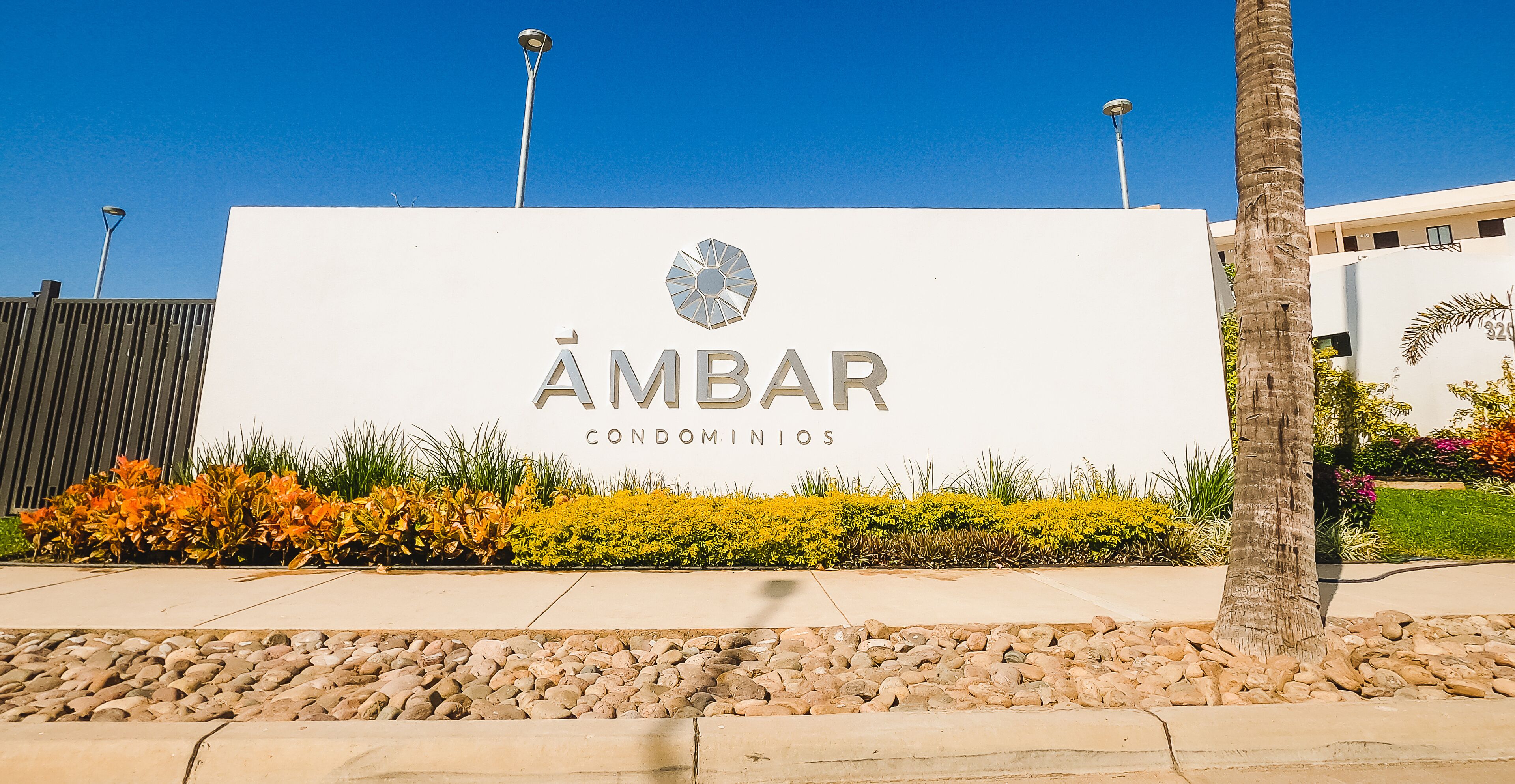 Señalización de 'ÁMBAR CONDOMINIOS' con un logotipo, rodeada de un área ajardinada con rocas y plantas.