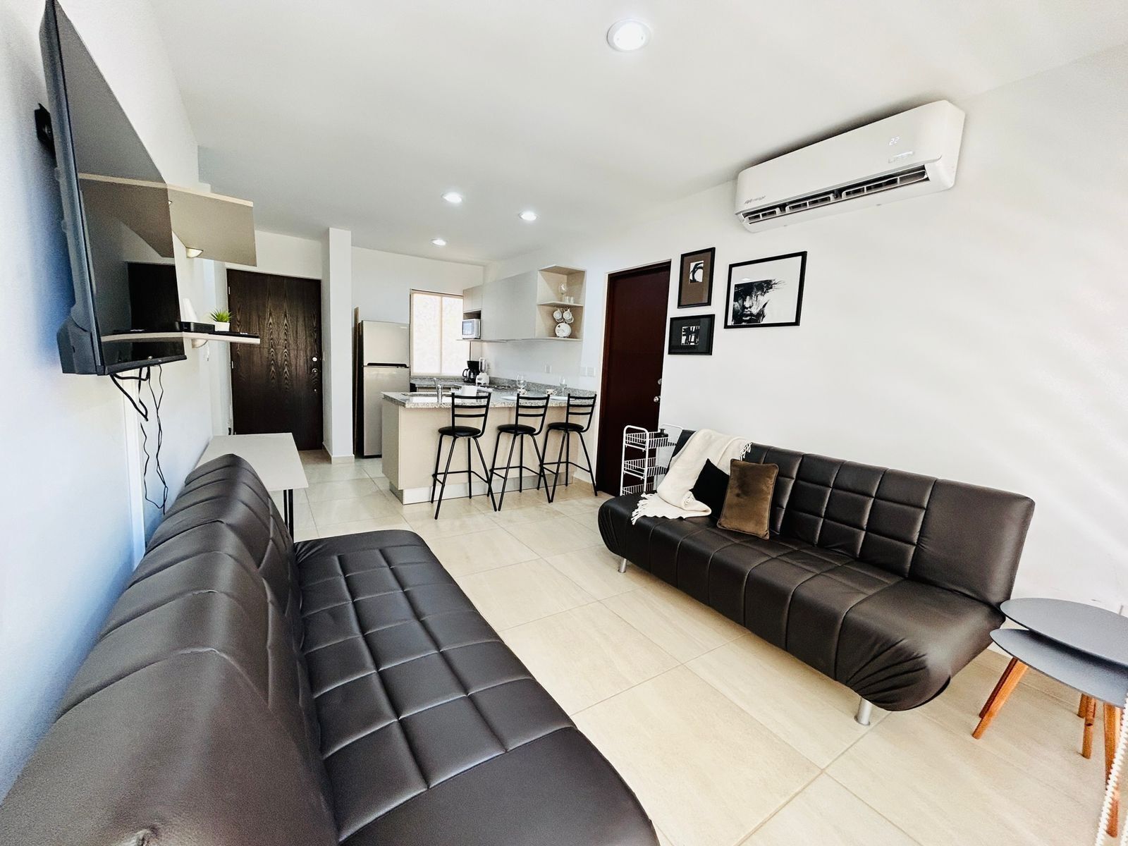 Una sala de estar moderna con un sofá de cuero negro, una mesita auxiliar y una cocina al fondo.