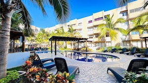 A resort pool area with lounge chairs, palm trees, and a building with balconies. - Ambar Apartments (Mazatlán)