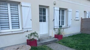 A house with a front door, two windows with shutters, a small garden with a table, and potted plants.