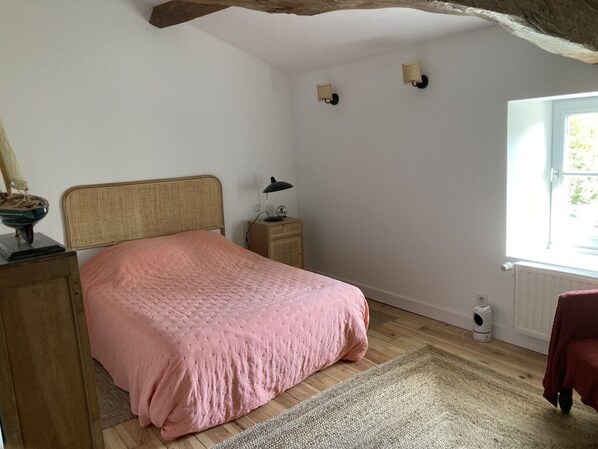 Un dormitorio con cabecera de madera, colcha rosa, mesita de noche con lámpara, radiador y ventana con vistas al verdor.