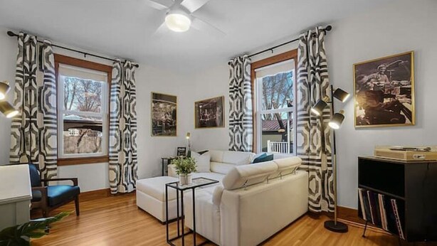 A modern living room with a sofa, a coffee table, and a bookshelf. There are large windows with curtains, a ceiling fan, and framed pictures on the wall. - Vitality Retreat - 1 block from DT/riverfront/shops/bars (Washington)