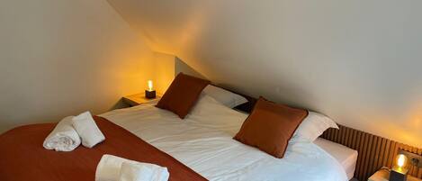 A neatly made bed with white linens and brown pillows, a red throw blanket, and two white towels on the bed. There is a nightstand with a lamp on it to the right of the bed.