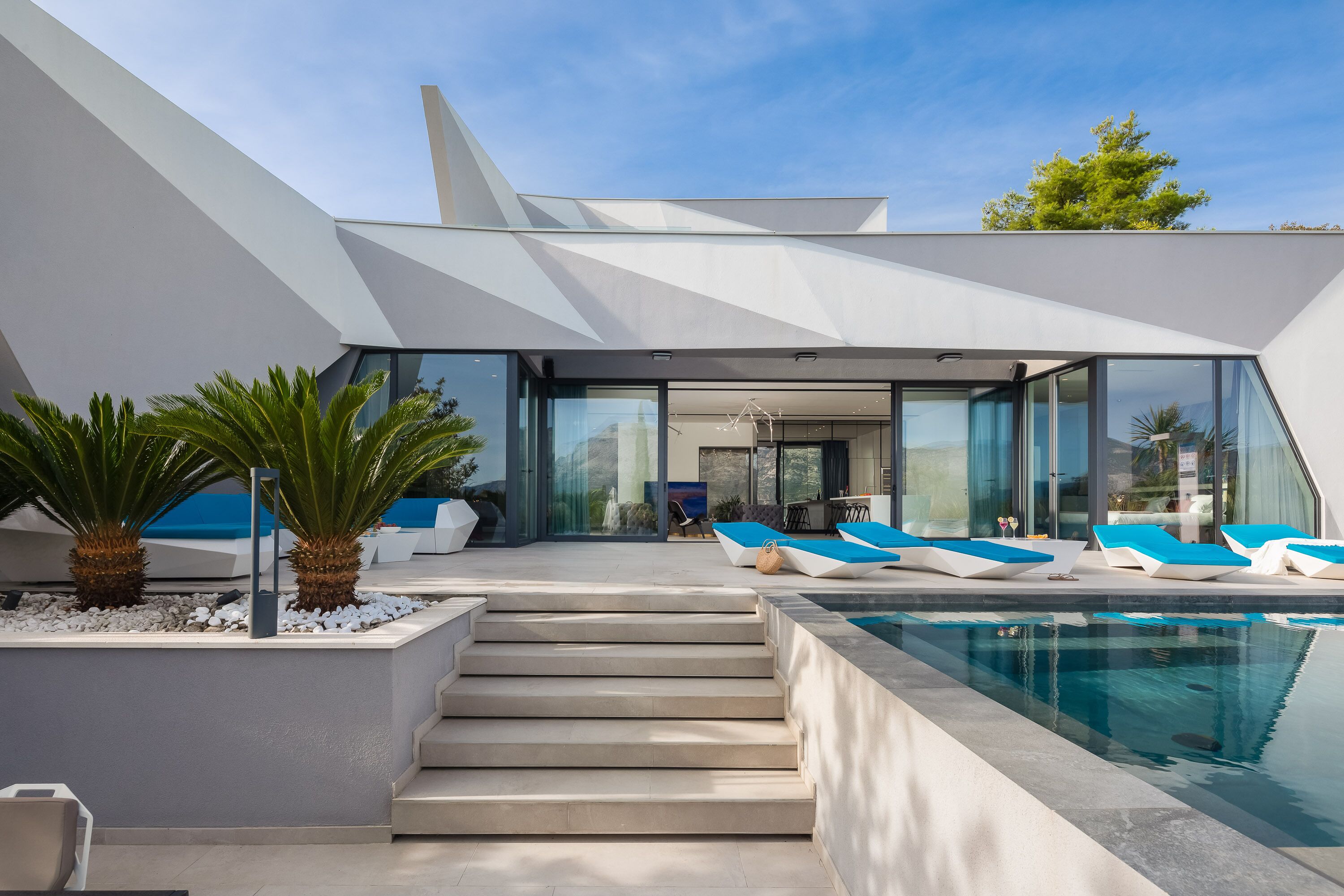 A modern outdoor pool area with a geometric poolside design, a staircase leading to a covered seating area, and a clear view of the sky.