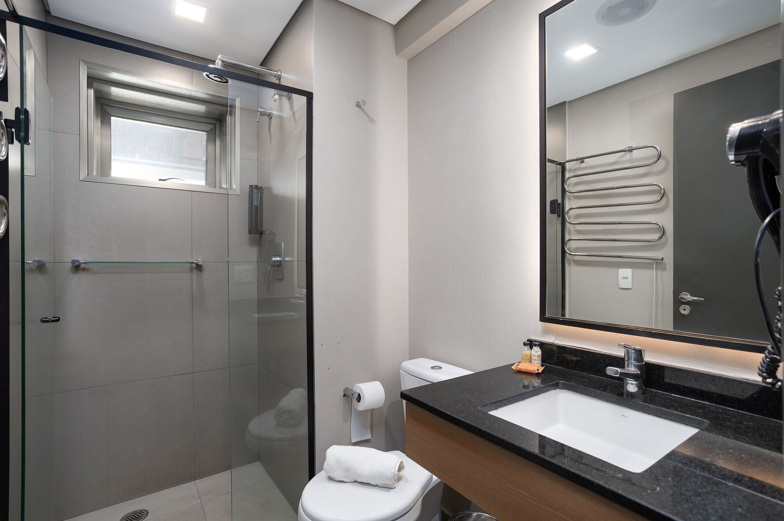 A modern bathroom with a glass shower enclosure, a white toilet, a sink with a black countertop, and a mirror with a towel rack.