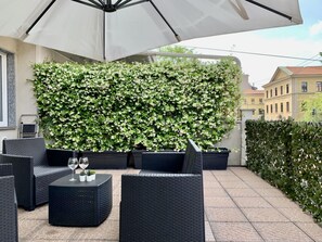 An outdoor seating area with wicker furniture, a white umbrella, and a wall of flowering plants. - Bright terrace in Milan (Milano)