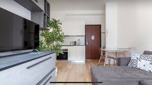 A modern living room with a grey sofa, a TV, a dining table with chairs, a kitchen area with cabinets, and a large plant. - Bright terrace in Milan (Milano)
