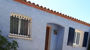 Une maison à l’extérieur bleu, avec deux tonneaux en bois et une fenêtre ornée de barreaux en métal.